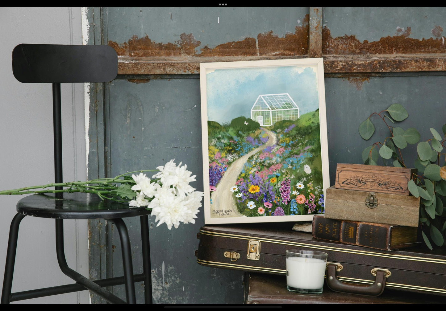 Framed artwork of a garden scene on a table with a black chair and decorative items.