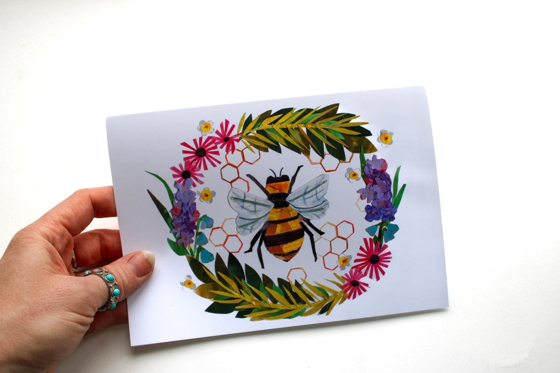 Hand holding a card with a colorful collaged bee illustration surrounded by flowers on a white background