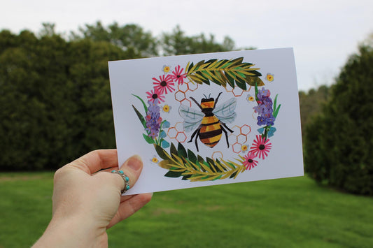 Hand holding a card with a paper collaged bee and floral design outdoors