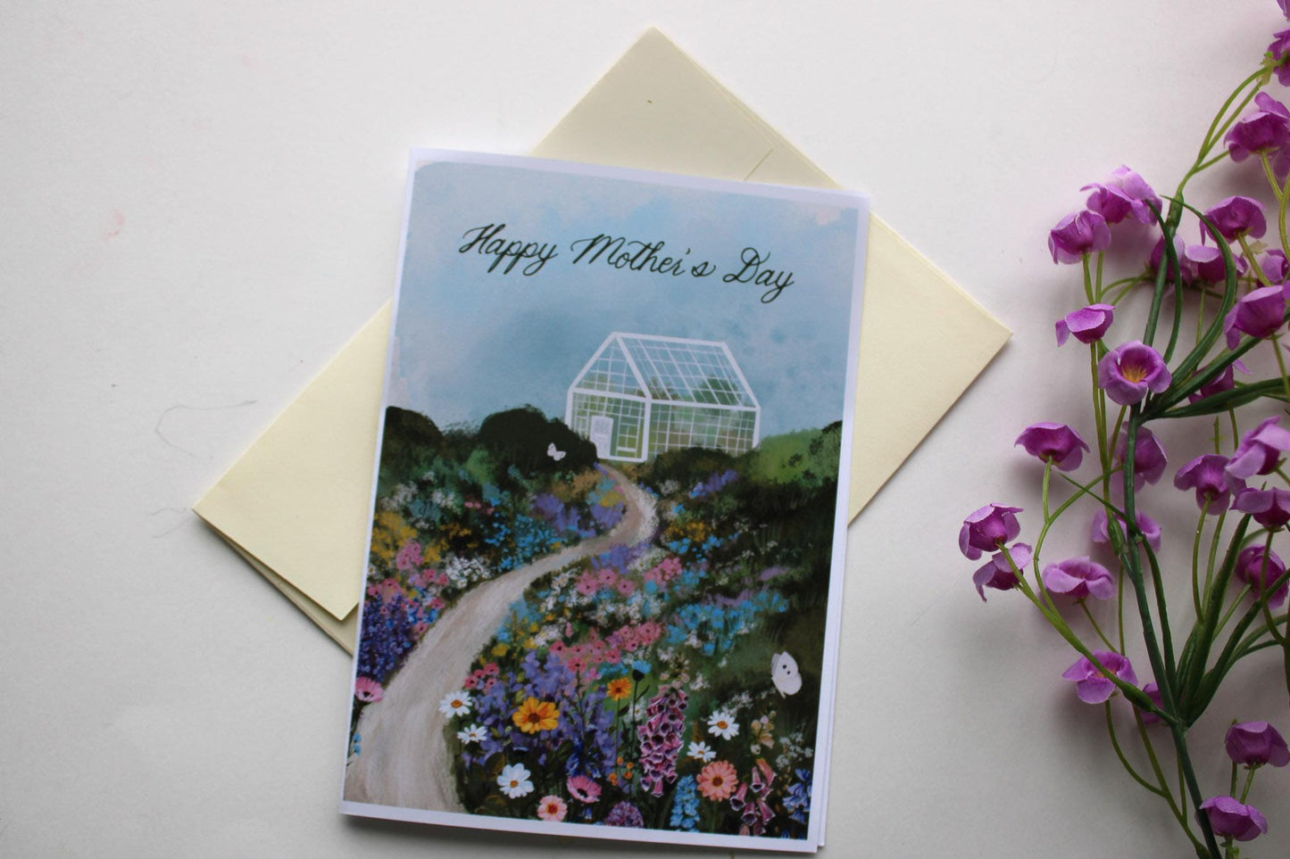Greeting card with 'Happy Mother's Day' on a garden design, next to a yellow envelope and purple flowers.