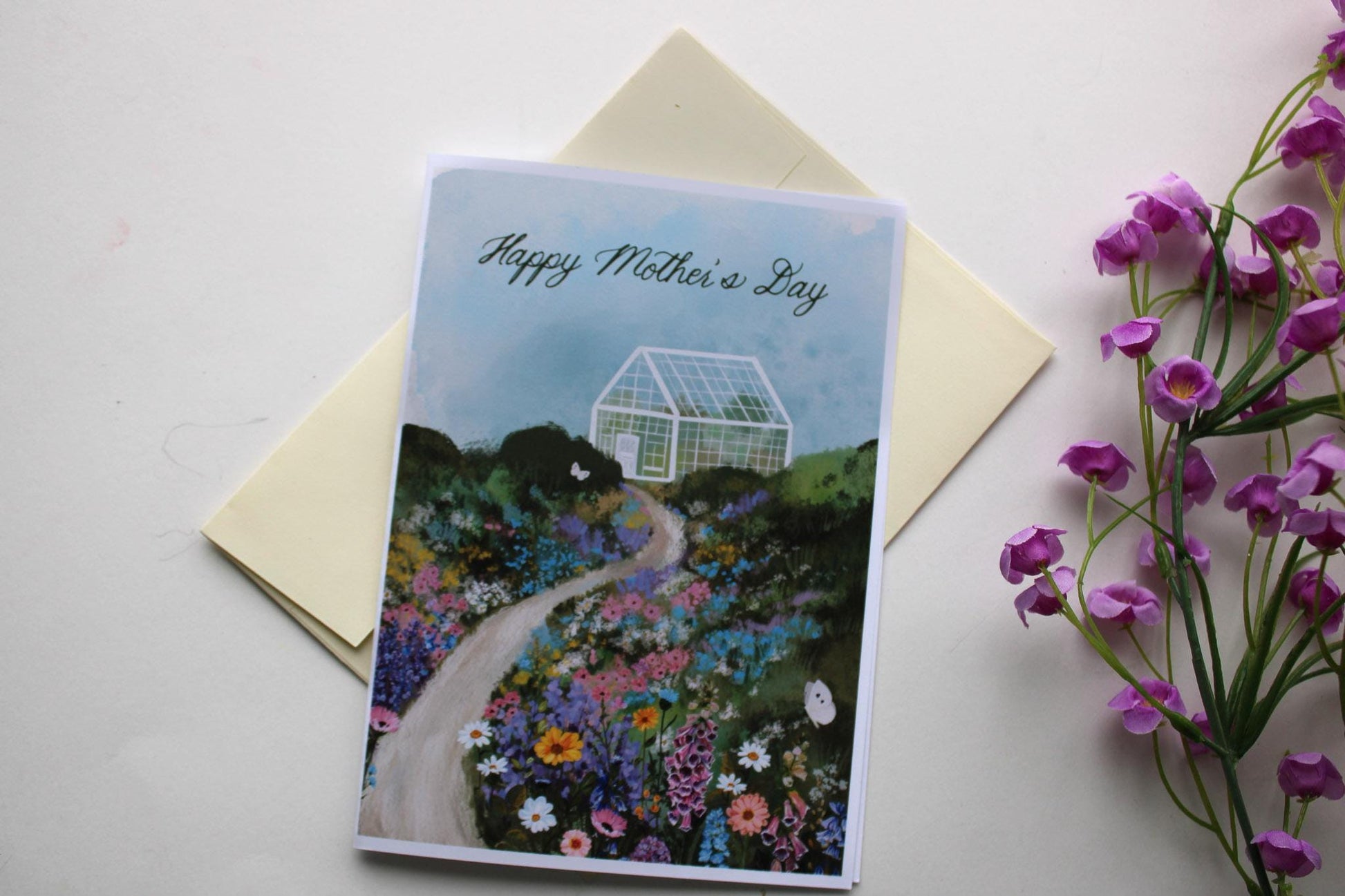 Greeting card with 'Happy Mother's Day' on a garden design, next to a yellow envelope and purple flowers.