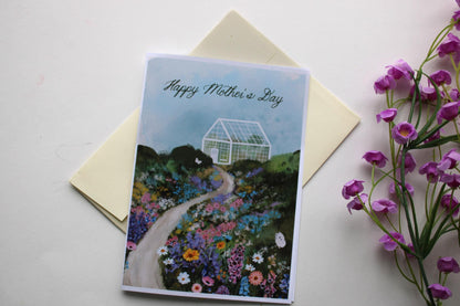 Greeting card with 'Happy Mother's Day' on a garden design, next to a yellow envelope and purple flowers.
