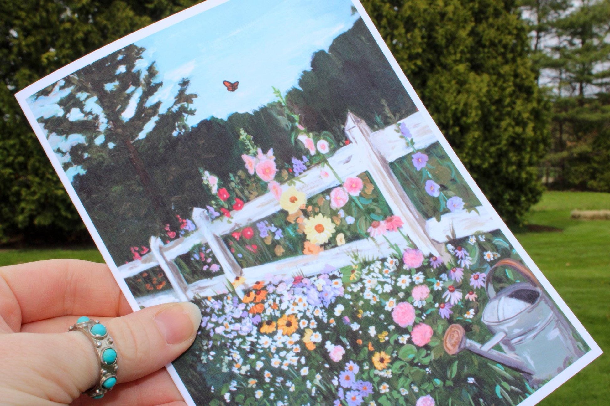 Hand holding a postcard with a garden scene featuring flowers and a white picket fence.