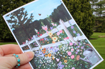Hand holding a postcard with a garden scene featuring flowers and a white picket fence.