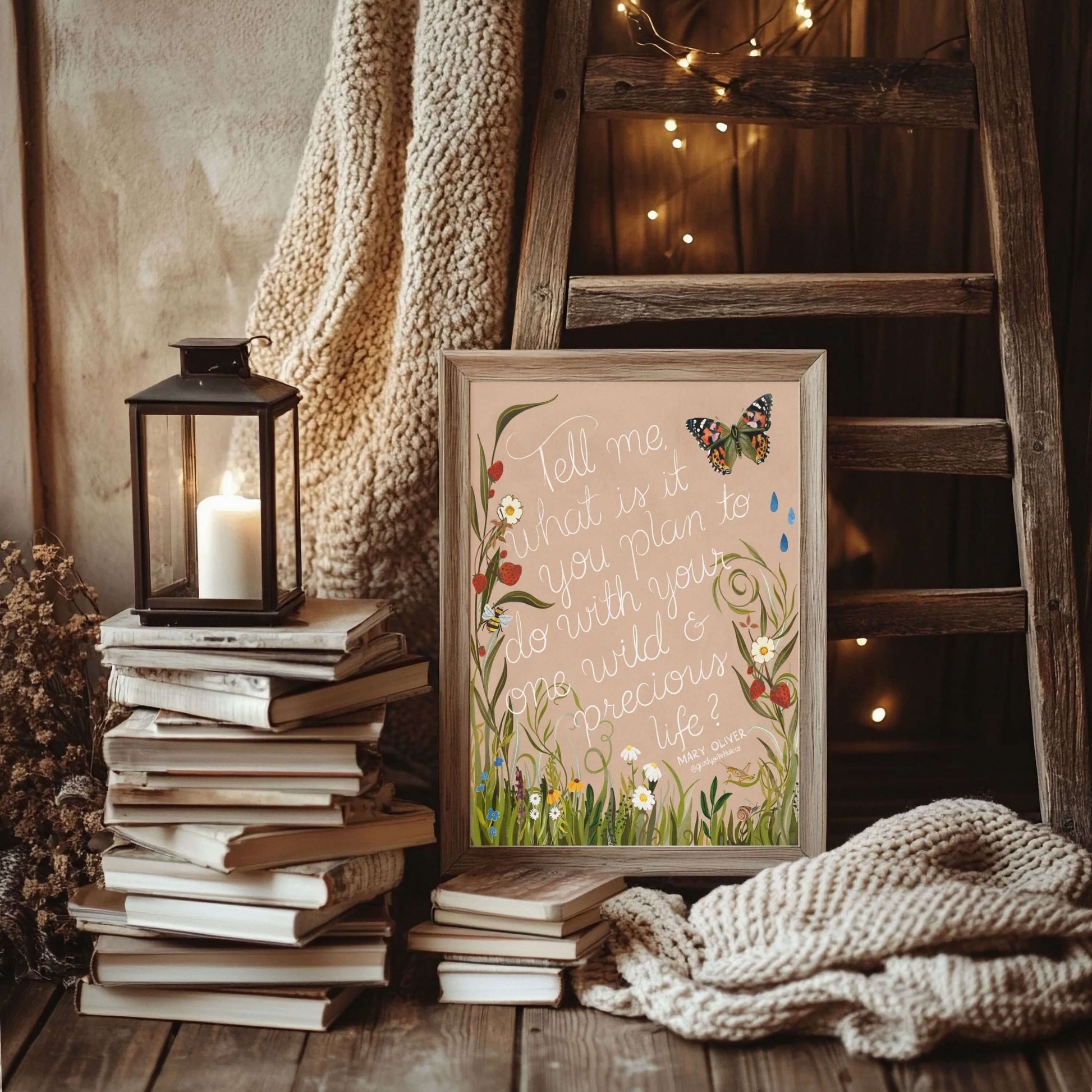 Framed artwork with floral design and butterfly, placed on a stack of books with a lantern and cozy blanket in a warm interior setting.