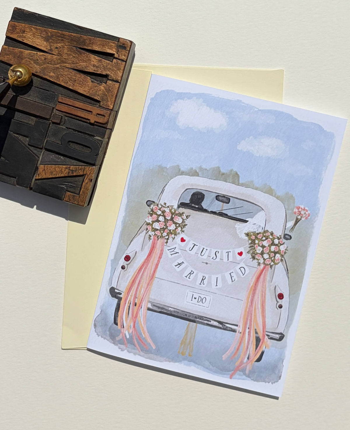 A square wedding greeting card featuring a watercolor illustration of a white vintage car with pink ribbons and flowers and the words 'Just Married' on the back, displayed on a cream envelope with a wooden letterpress block at the side.