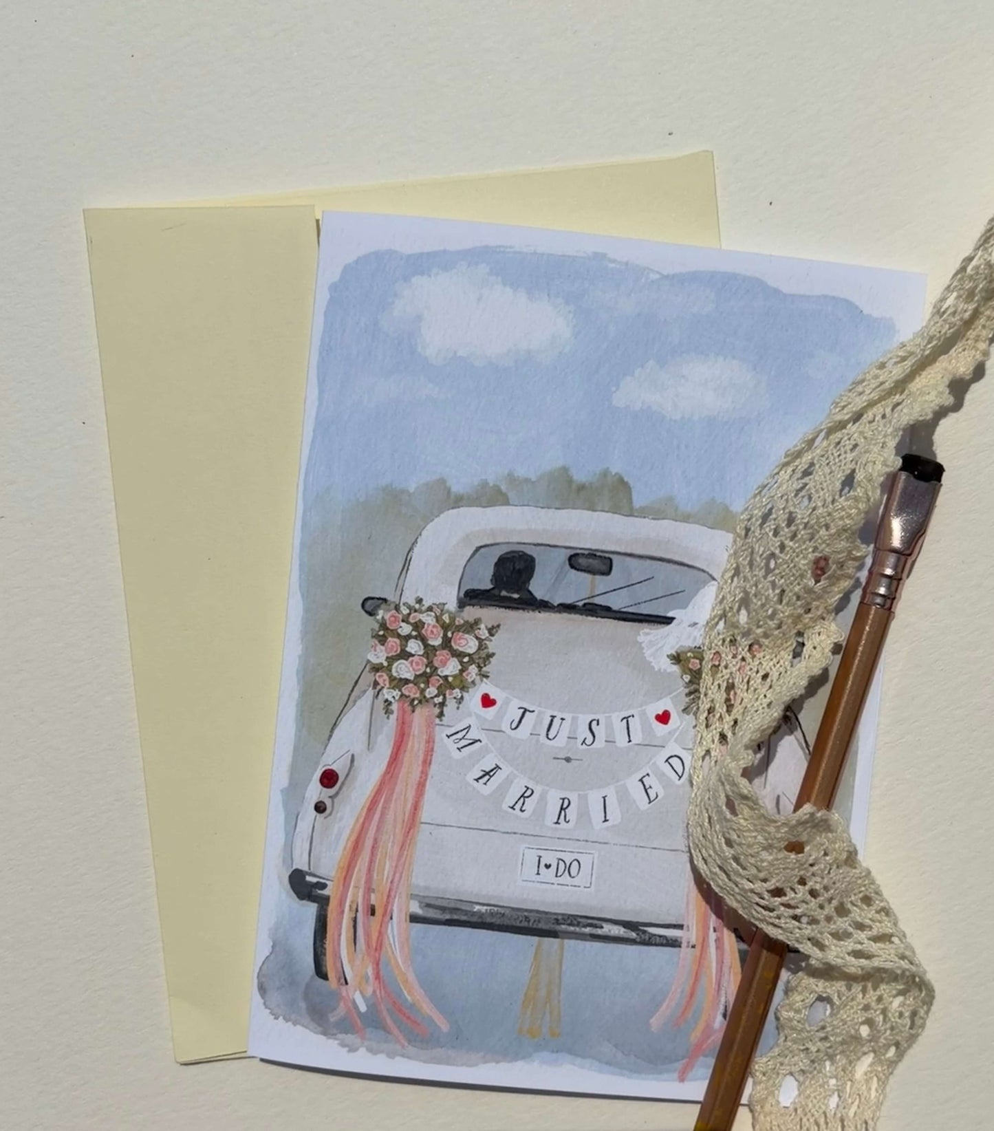 A wedding greeting card featuring a watercolor illustration of a white car with pink ribbons and flowers, alongside a cream envelope, a paintbrush, and a lace ribbon on a light beige background.