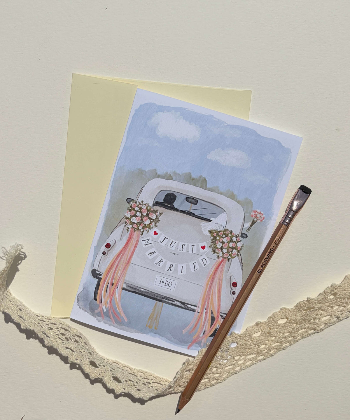 A watercolor-style greeting card featuring the back of a white wedding car with pink ribbons and flowers and the words 'Just Married' and '1990', displayed with a pale yellow envelope, a wooden paintbrush, and cream lace on a light beige surface.