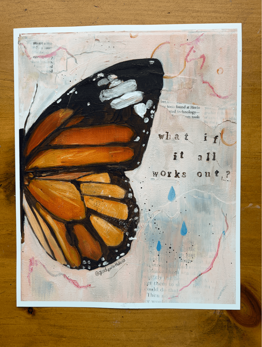 An art print featuring a painted monarch butterfly on a textured, collage-style background with the text 'what if it all works out?' and blue raindrop accents, displayed on a wooden surface.