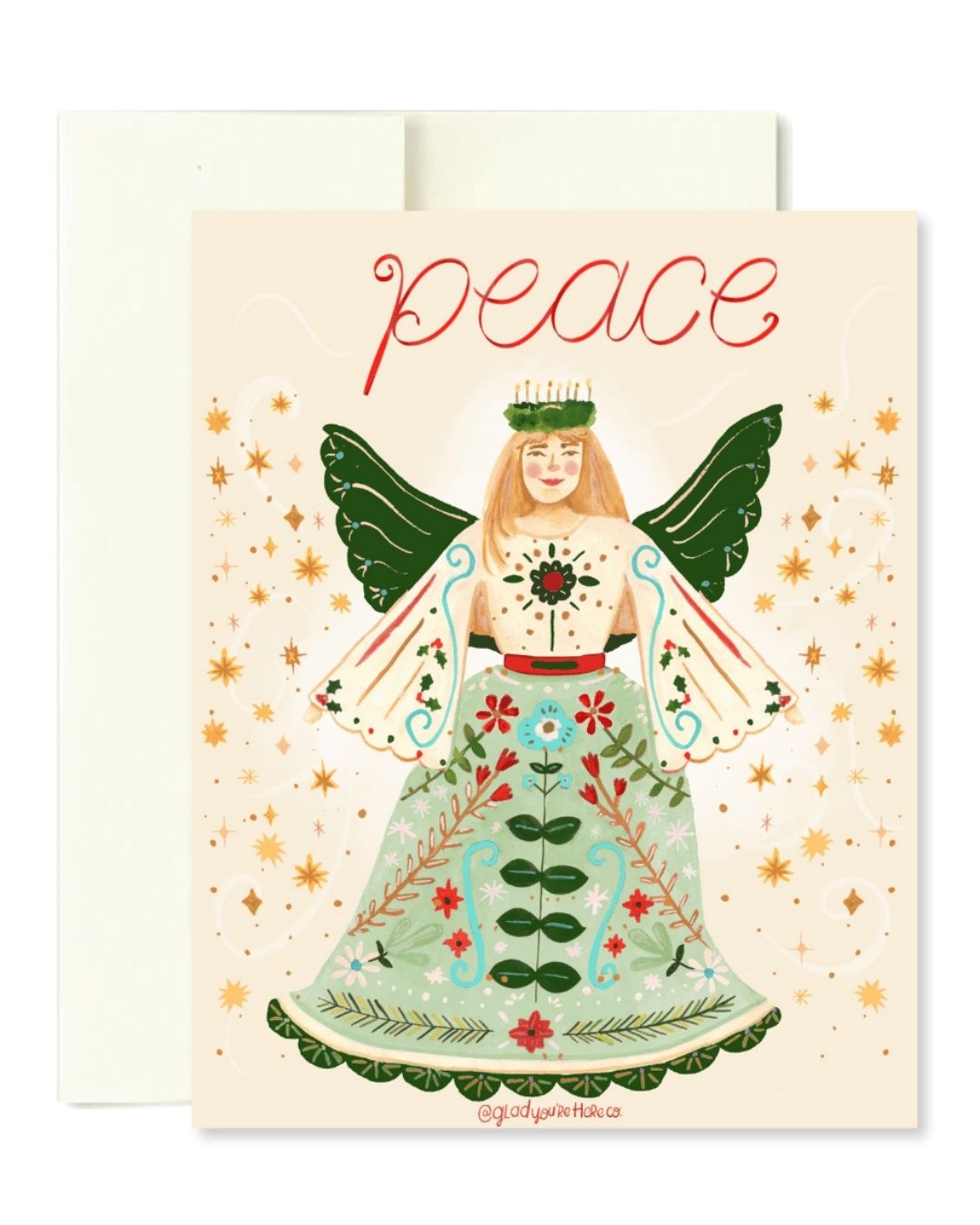A greeting card featuring an illustrated angel with green wings, a floral dress, and the word 'peace' in red script at the top, shown with a cream envelope behind it on a white background.