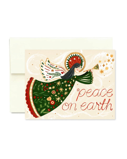 A Christmas greeting card with an illustrated angel in a green patterned dress blowing a trumpet, with the words 'peace on earth' in red script, shown with a cream envelope behind it on a white background.