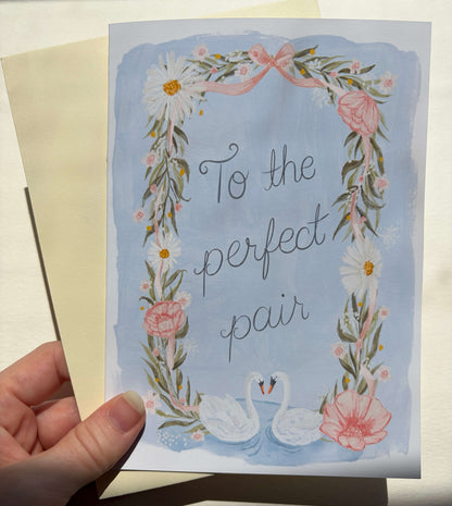 A hand holds a square greeting card with a watercolor-style floral wreath and two swans, with the text 'To the perfect pair.' A cream envelope is partially visible behind the card.