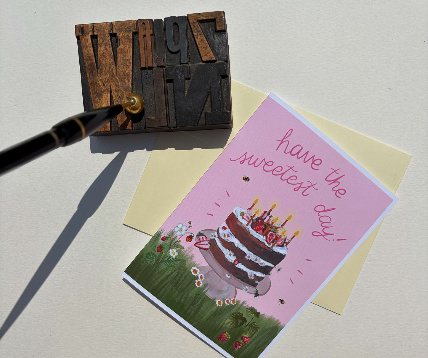 A pink birthday card with an illustration of a layered cake and the words 'have the sweetest day!' is displayed on a cream envelope, next to a wooden letterpress block and a black pen on a light beige background.