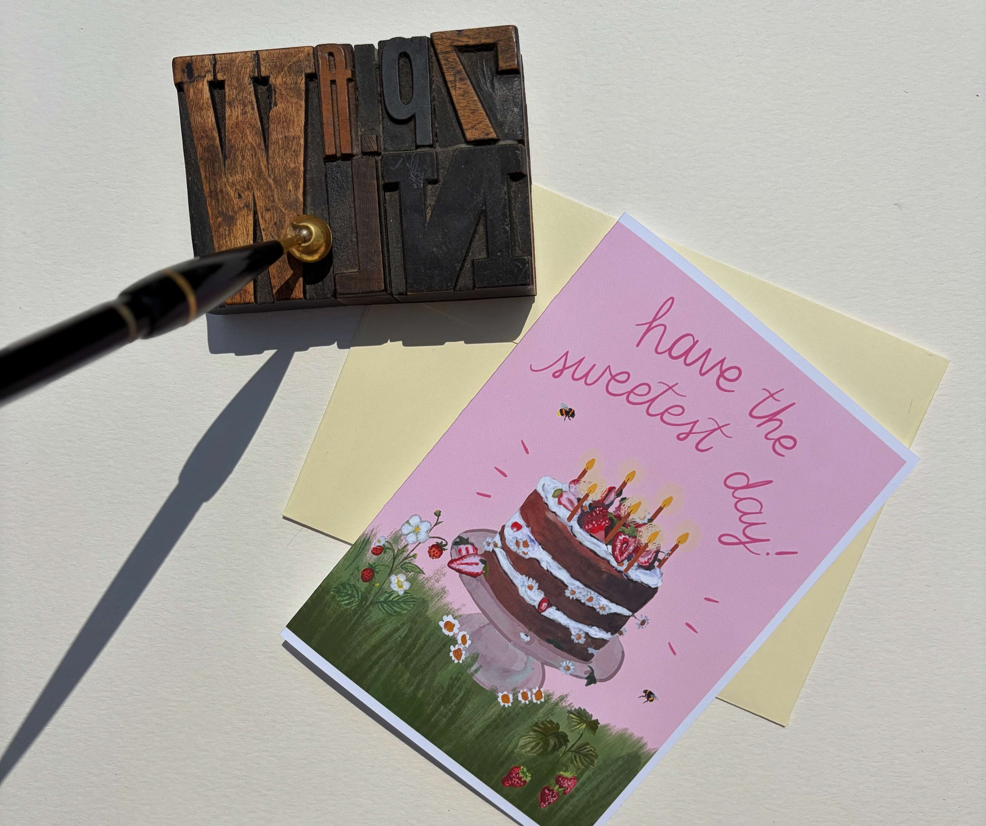 A pink birthday card with an illustration of a layered cake and the words 'have the sweetest day!' is displayed on a cream envelope, next to a wooden letterpress block and a black pen on a light beige background.