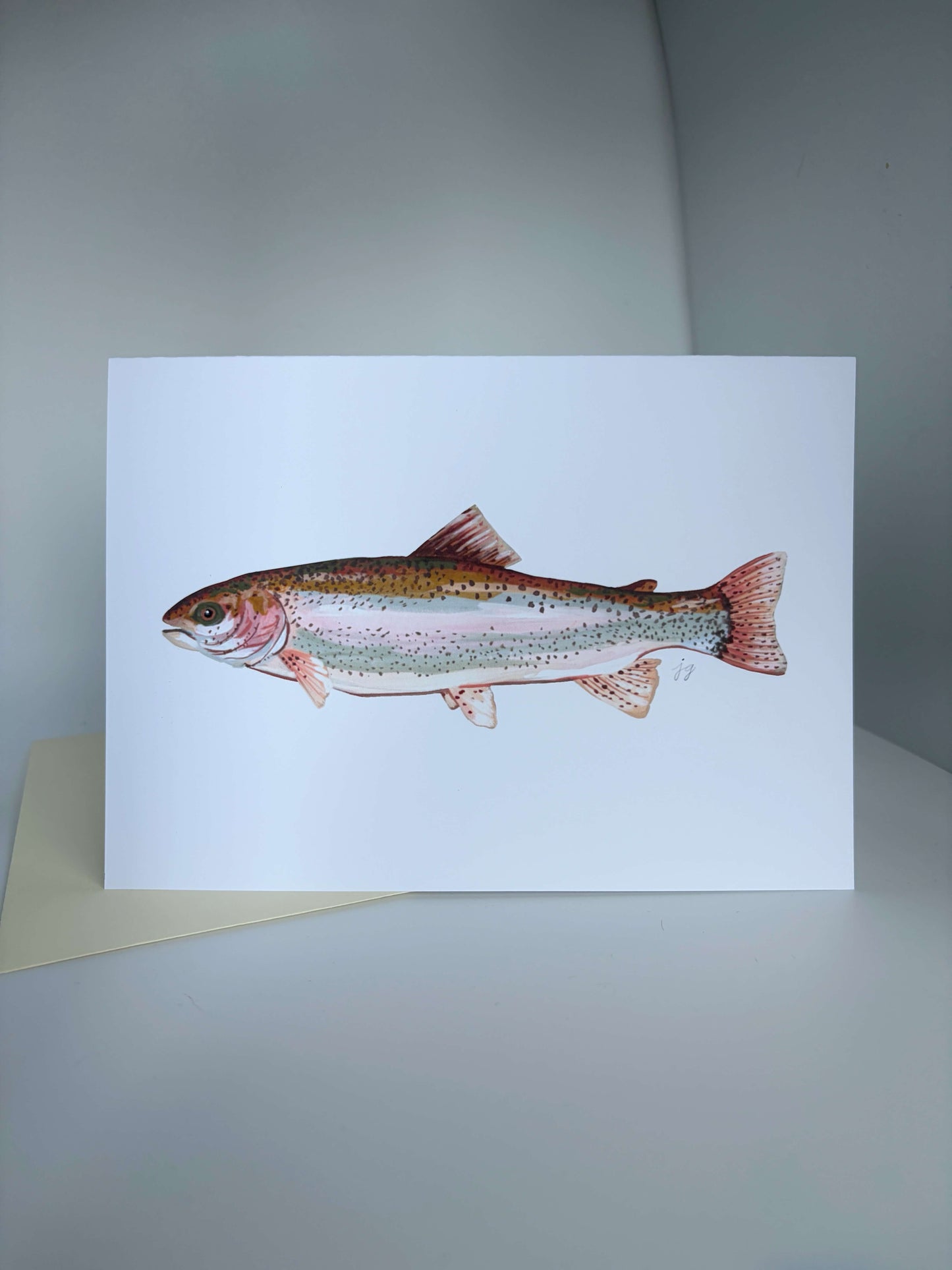 A white greeting card featuring a detailed watercolor illustration of a rainbow trout, displayed on a neutral gray surface with a beige envelope partially visible underneath.
