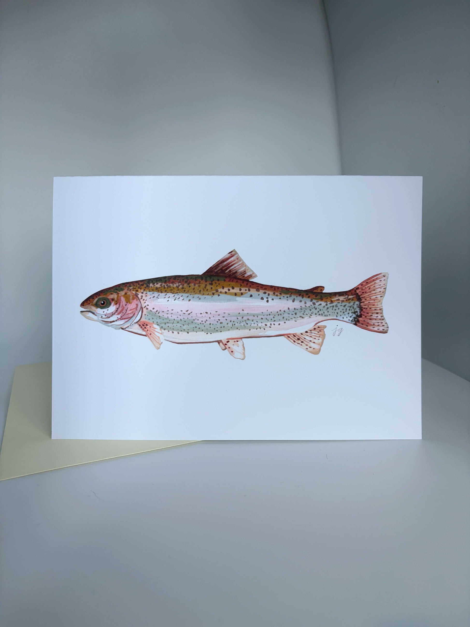 A white greeting card featuring a detailed watercolor illustration of a rainbow trout, displayed on a neutral gray surface with a beige envelope partially visible underneath.