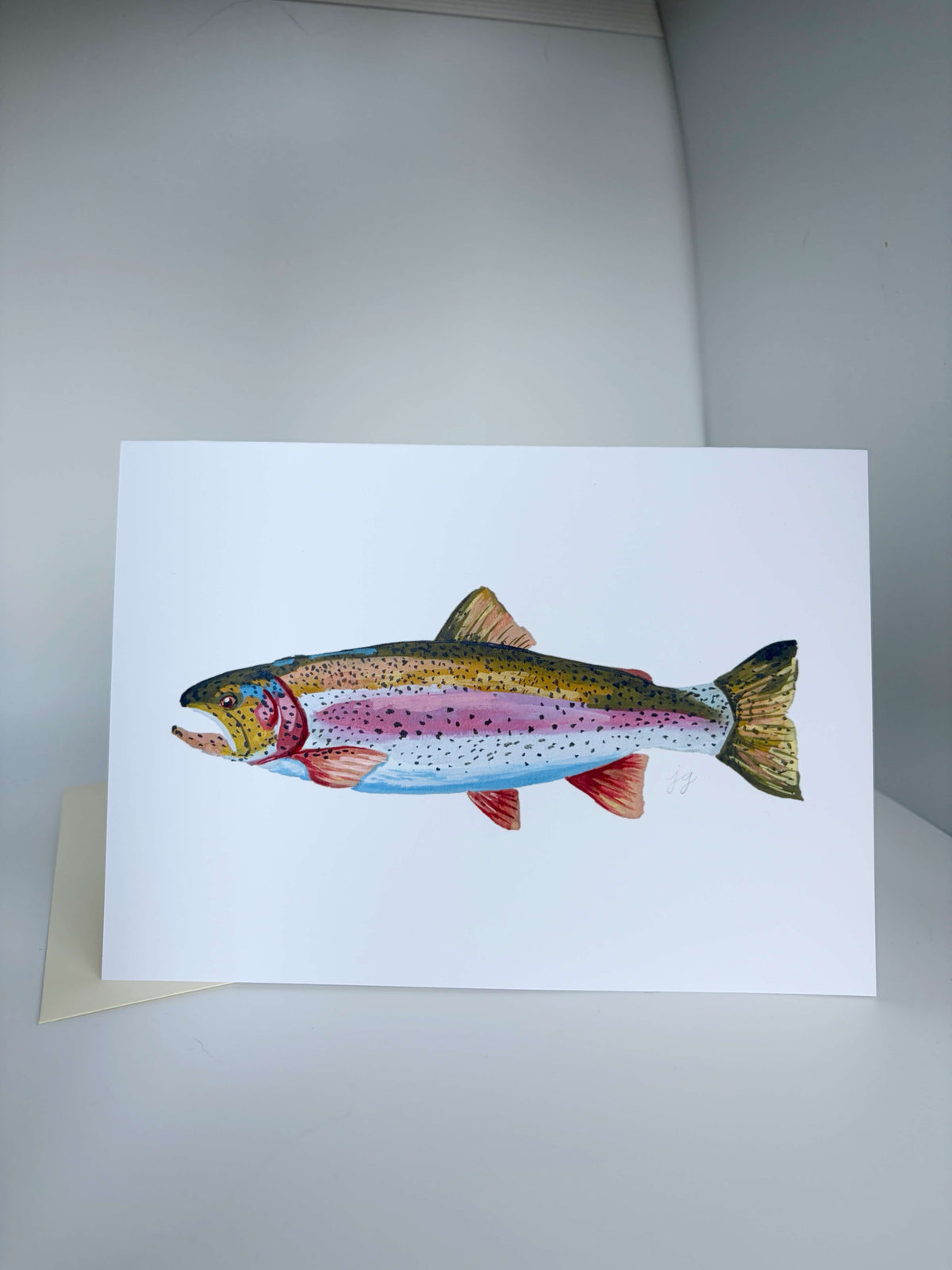 A white art print card featuring a detailed watercolor illustration of a rainbow trout, displayed on a neutral gray surface with a small kraft envelope partially visible underneath.