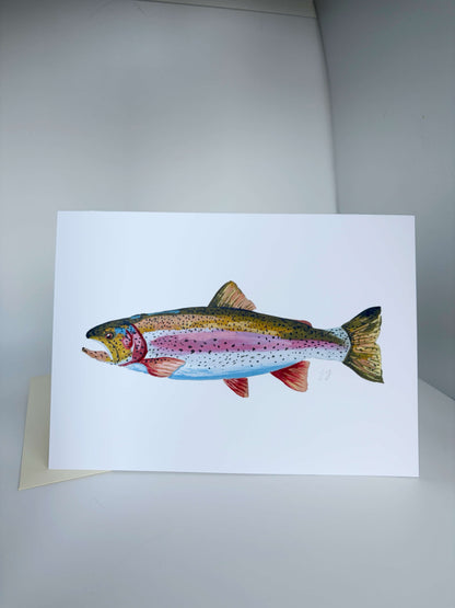 A white art print card featuring a detailed watercolor illustration of a rainbow trout, displayed on a neutral gray surface with a small kraft envelope partially visible underneath.