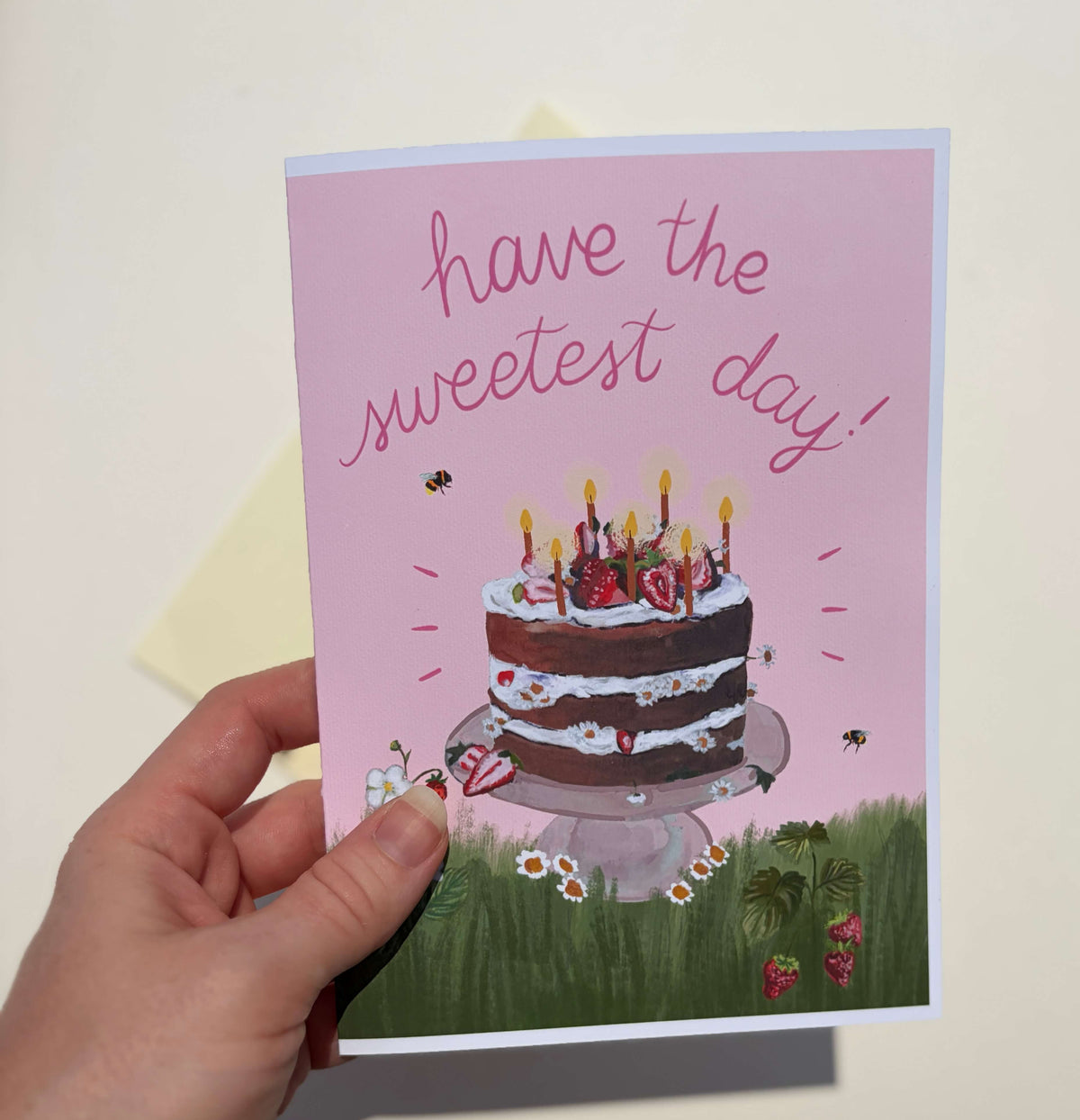 A hand holding a pink greeting card that reads 'have the sweetest day!' with an illustration of a birthday cake, candles, bees, and grass at the bottom. A pale envelope is visible behind the card against a light neutral background.
