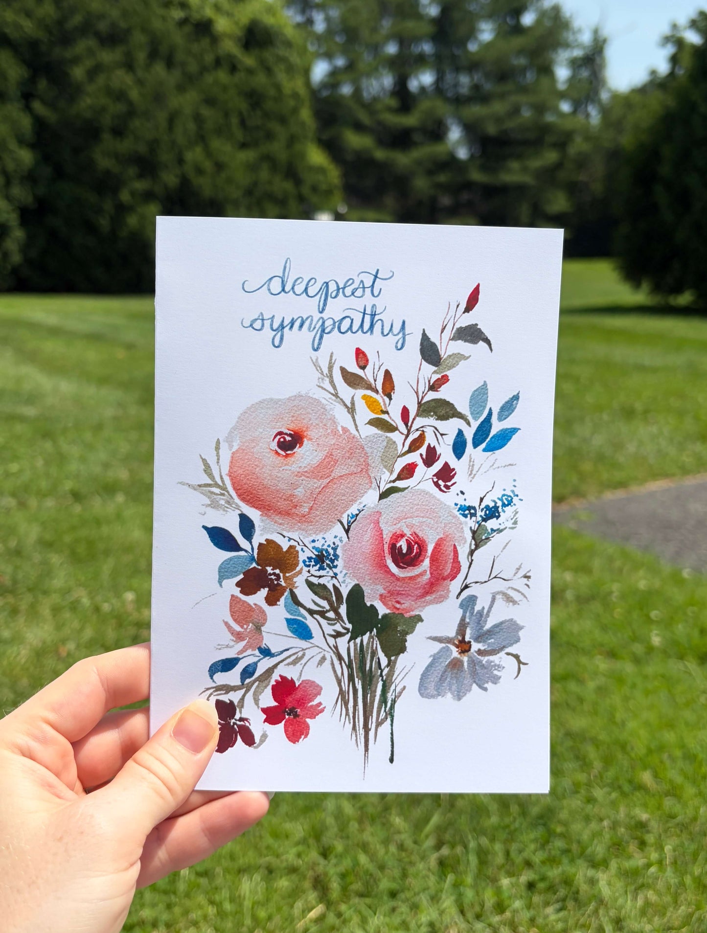 A hand holds a white greeting card with a watercolor floral bouquet and the words 'deepest sympathy' in blue script, photographed outdoors against a blurred green lawn and trees.