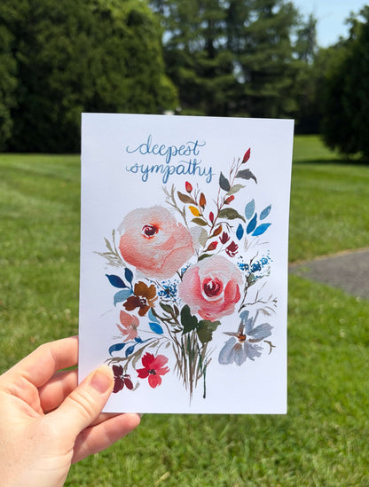 A hand holds a white greeting card with a watercolor floral bouquet and the words 'deepest sympathy' in blue script, photographed outdoors against a blurred green lawn and trees.