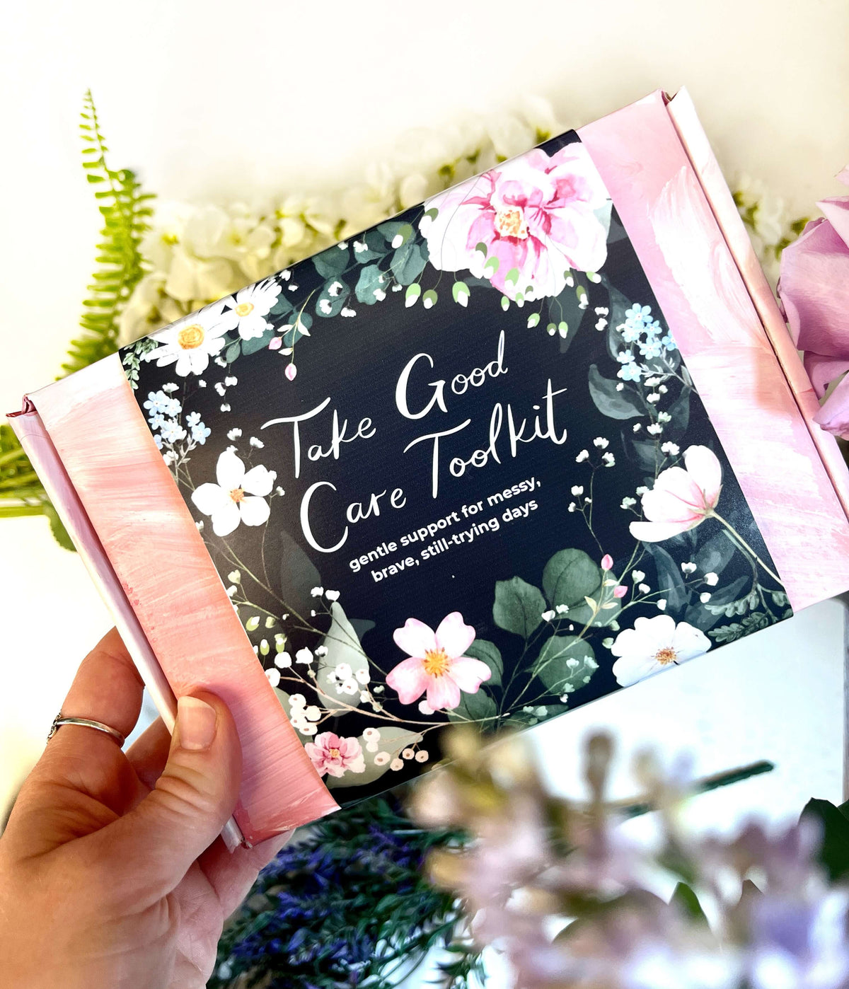 Hand holding a 'Take Good Care Toolkit' with floral design and text.