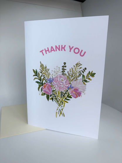 A white greeting card standing upright on a white surface, printed with the words 'THANK YOU' in pink above a watercolor-style bouquet of pink, purple, green, and cream flowers. A cream envelope is partially visible underneath the card.