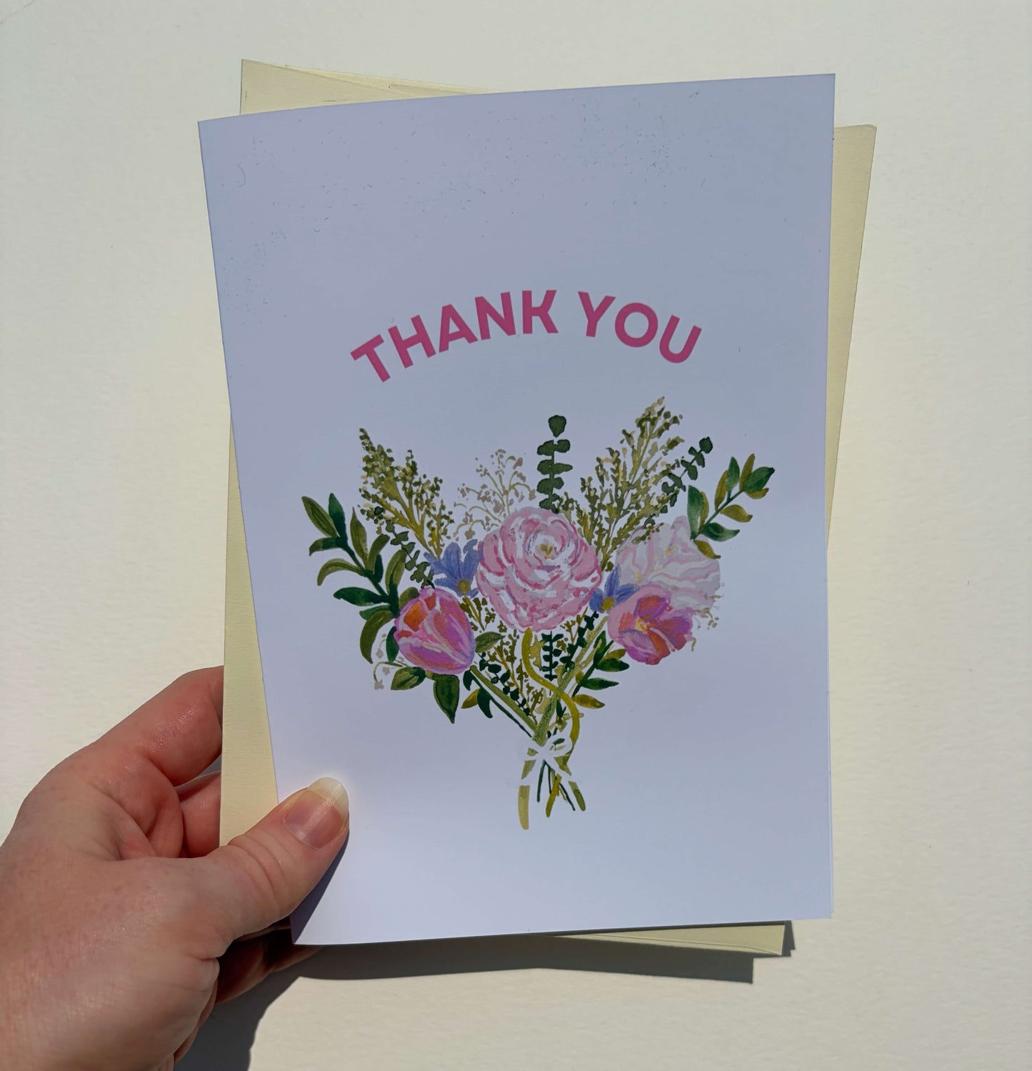 A hand holding a square thank you card with a watercolor-style bouquet of pink and purple flowers and green leaves, with the words 'THANK YOU' in pink at the top. A cream envelope is partially visible behind the card against a light beige background.