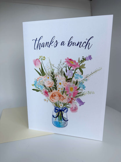 A white greeting card standing upright on a light surface, printed with the phrase 'thanks a bunch' in dark script above a watercolor-style bouquet of pink, peach, blue, purple, and green flowers in a blue glass vase. A cream envelope is partially visible underneath the card.