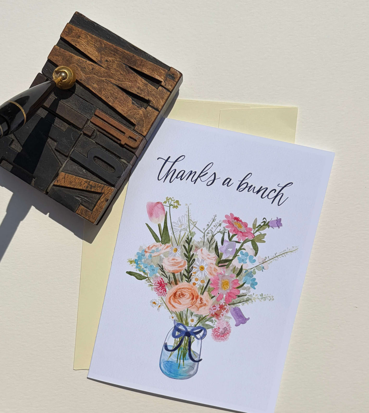 A white greeting card with the phrase 'thanks a bunch' above a watercolor-style bouquet of pink, peach, blue, and purple flowers in a blue vase, styled on a cream surface with a pale yellow envelope and a dark wooden stamp block.