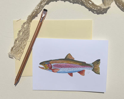 A watercolor-style trout illustration printed on a white card, styled on a cream surface with a pencil, pale yellow paper, and a piece of lace.