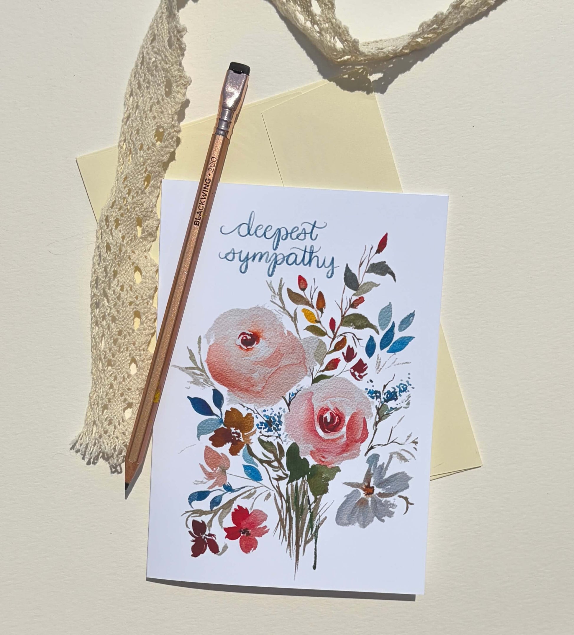 A white greeting card with a watercolor floral bouquet and the words 'deepest sympathy' in blue script, styled on a cream background with a yellow envelope, a wooden paintbrush, lace trim, and a rope.