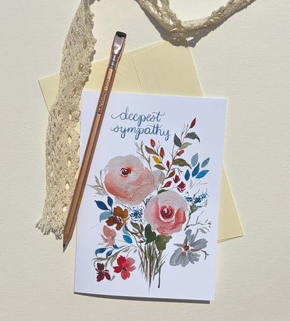 A white greeting card with a watercolor floral bouquet and the words 'deepest sympathy' in blue script, styled on a cream background with a yellow envelope, a wooden paintbrush, lace trim, and a rope.