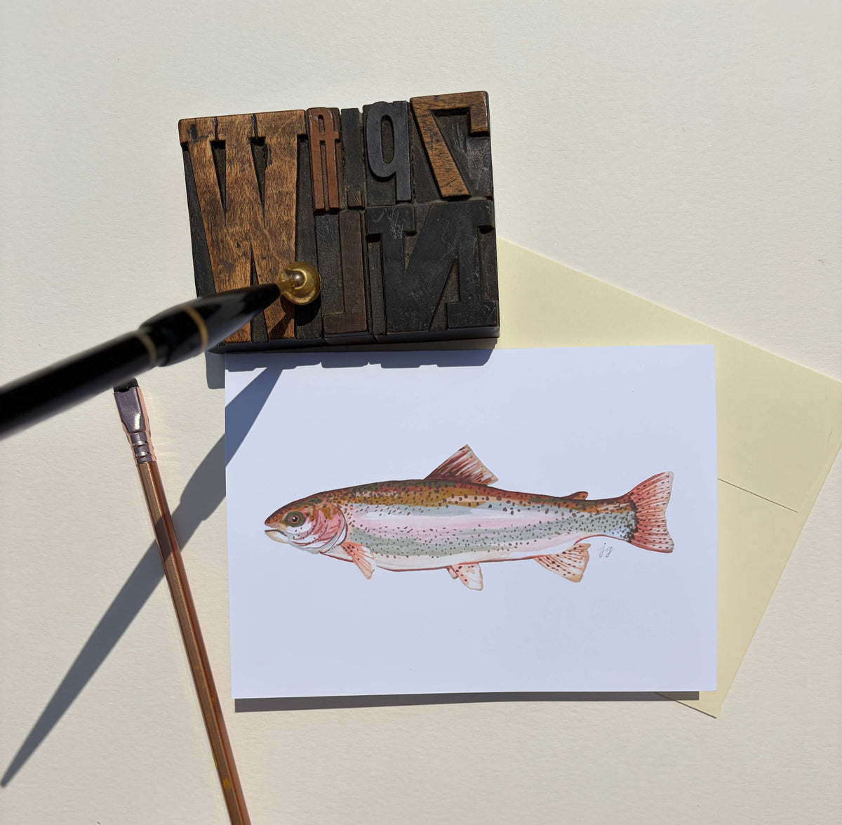 A white art print card featuring a detailed watercolor illustration of a rainbow trout, styled on a cream background with a wooden letterpress block, a paintbrush, and a paint tube.