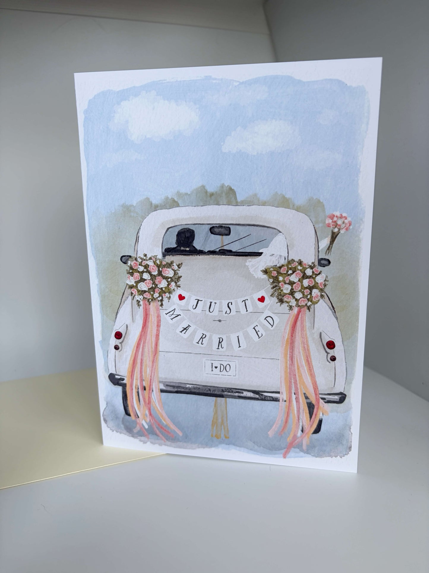A vertical greeting card featuring a watercolor illustration of a white vintage car decorated with floral bouquets and pink ribbons, with the words 'JUST MARRIED' across the back. The card is photographed standing upright on a light surface against a neutral gray background.