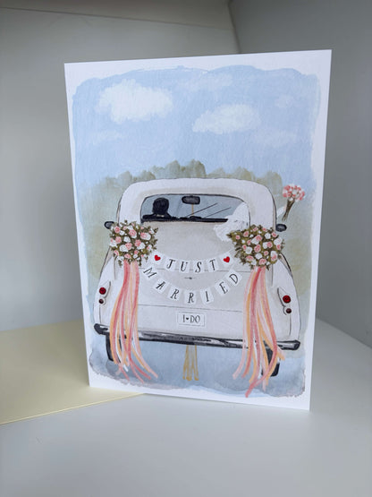 A vertical greeting card featuring a watercolor illustration of a white vintage car decorated with floral bouquets and pink ribbons, with the words 'JUST MARRIED' across the back. The card is photographed standing upright on a light surface against a neutral gray background.