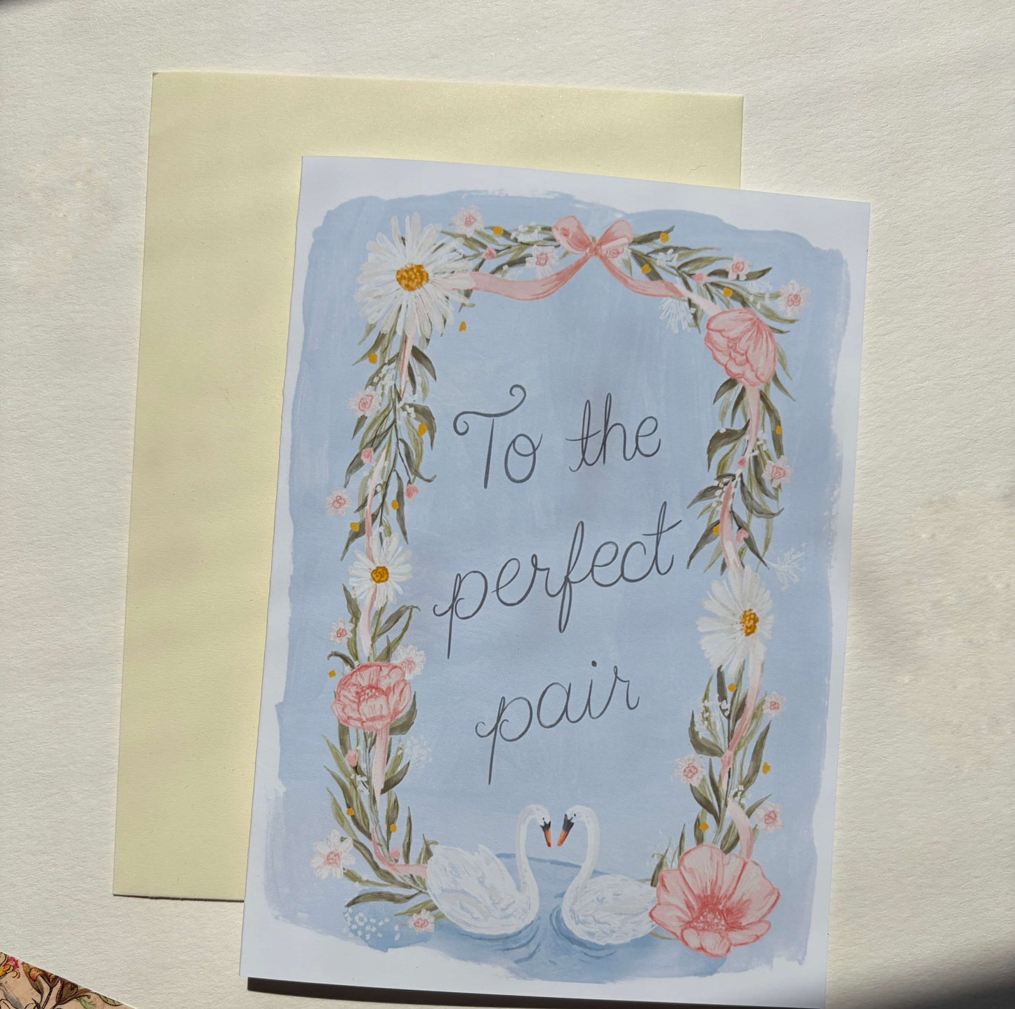 A 5x7 greeting card with a floral wreath border, two swans at the bottom, and the phrase 'To the perfect pair' in script, placed in front of a cream envelope on a light textured surface.