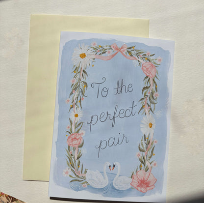A 5x7 greeting card with a floral wreath border, two swans at the bottom, and the phrase 'To the perfect pair' in script, placed in front of a cream envelope on a light textured surface.